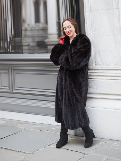 Woman leaning against wall in dark ranch black mink fur coat. Timeless fur fashion with cozy and elegant appeal.