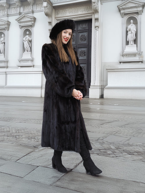 Model in black mink fur coat standing in front of ornate architecture. Full-length coat with luxurious finish and timeless appeal.