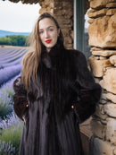 Woman in a dark fur coat standing in front of a stone wall with a scenic background.
