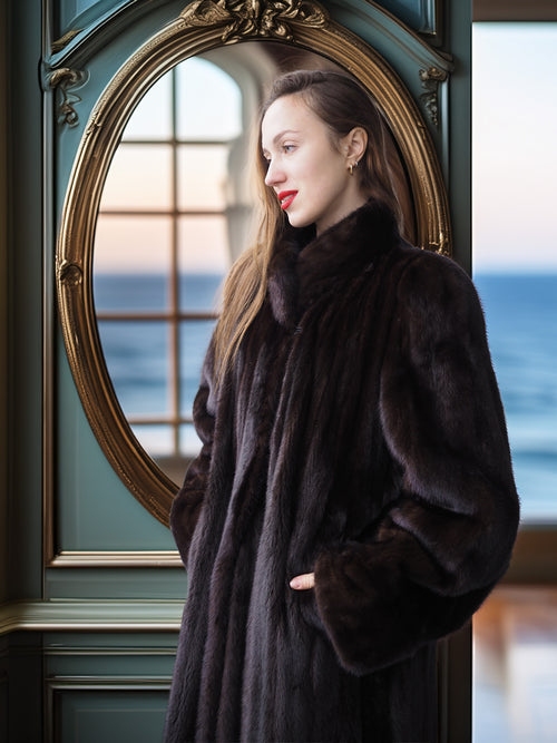 Woman in a fur coat standing in front of a large mirror with a scenic view.