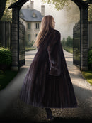 Woman in a long coat walking towards a grand house through an archway.