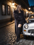 Woman in a long coat standing next to a vintage car on a cobbled street at night.