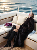 Woman in a fur coat and knee-high boots sitting on a boat with a plate of grapes.
