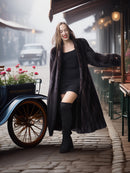 Woman in a long coat and black outfit standing in front of a vintage car on a street.