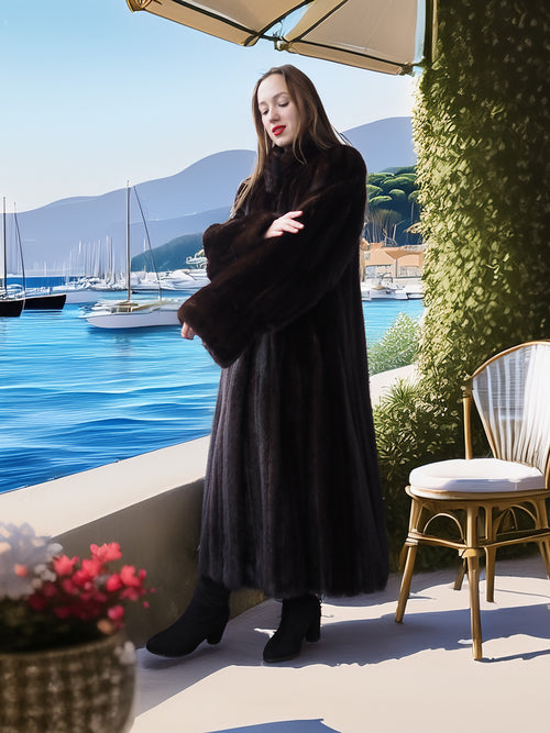 Woman in a long coat standing by a waterfront with sailboats and mountains in the background.