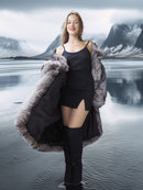 Woman in a long fur coat standing in front of a lake with mountains in the background