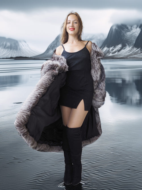 Woman in a long fur coat standing in front of a lake with mountains in the background