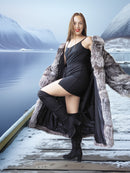 Woman in a black dress and fur coat standing on a wooden dock with mountains and water in the background