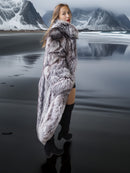 Woman wearing a fur coat standing in front of a mountainous landscape with water.