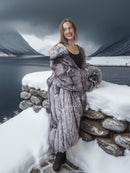 Woman in a fur coat standing on a snowy ledge with mountains in the background