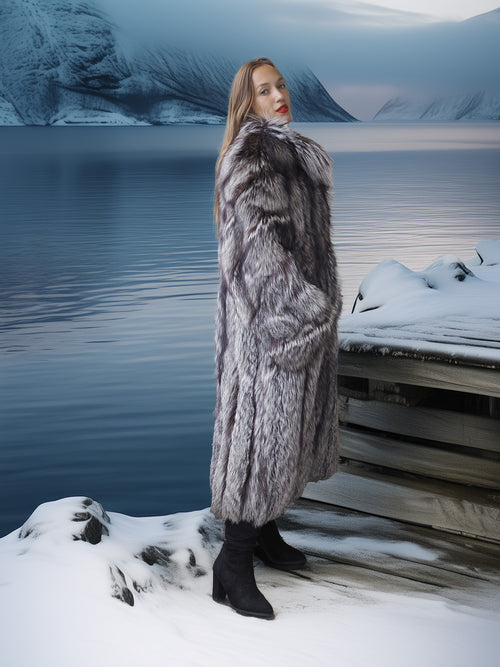 Person wearing a fur coat standing by a snowy landscape with mountains and water.