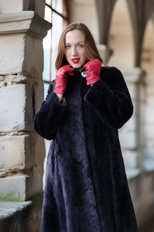 Woman wearing a long black coat and red gloves standing against a stone wall.