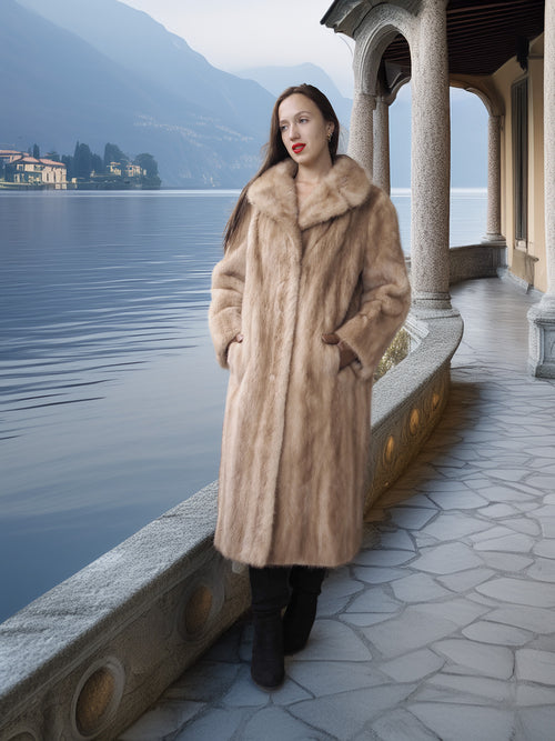 Woman in a fur coat standing by a lakeside with mountains in the background