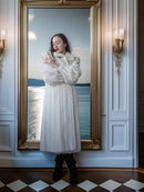 Woman in a white fur coat standing in front of a large mirror with a scenic view.