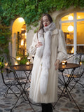 Woman in a long white fur coat standing in an outdoor setting with a building and chairs in the background.