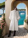 Woman in a white fur coat standing by a scenic waterfront with boats and flowers.