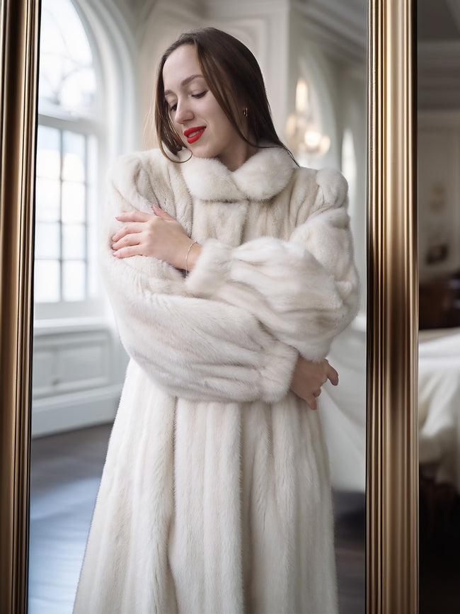 Woman wearing a white fur coat standing in front of a mirror.