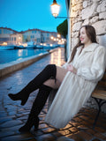Woman in a white coat sitting on a bench by a waterfront at dusk.