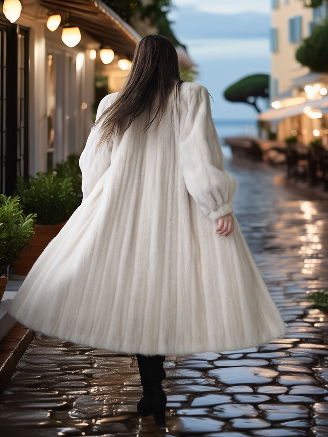 Person wearing a long white coat walking on a stone pavement with buildings and lights in the background.