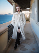 Woman in a white coat standing on a balcony with a scenic ocean view.