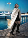 Woman in a white fur coat and black boots standing by a waterfront at dusk.