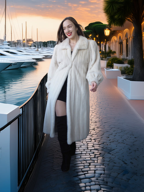 Woman in a long white coat walking along a waterfront promenade with yachts in the background.