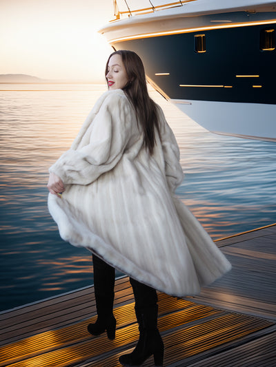Woman in a white coat standing on a dock with a yacht in the background
