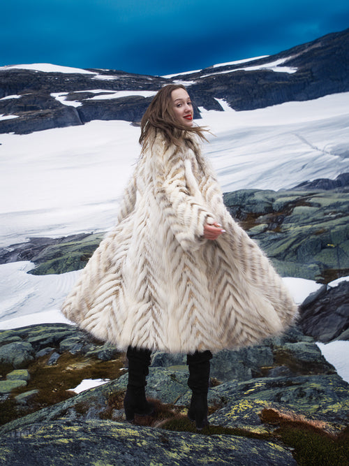Model twirling in Two Tone Fox Coat Size XS/S on rocky terrain, showcasing the coat’s dramatic chevron fur and winter elegance.