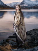 Woman in a fur coat standing by a lake with mountains in the background