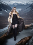 Woman in a fur coat and black dress standing in a snowy landscape with mountains.