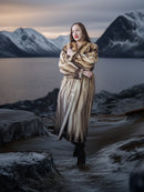 Woman in a fur coat standing on a rocky shoreline with mountains and water in the background
