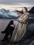 Woman in a fur coat sitting by a lake with mountains in the background