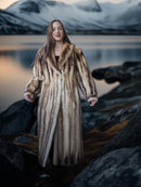 Woman in a fur coat standing by a lake with mountains in the background