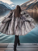 Person wearing a large fur coat standing on a dock with mountains and water in the background