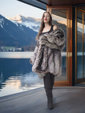 Woman in a fur coat standing in front of a large window with a mountain view.