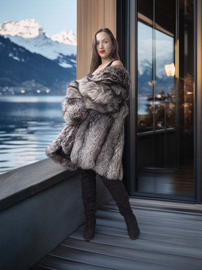 Woman in a fur coat standing by a window with a mountainous landscape view.
