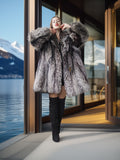 Woman in a fur coat standing by a large window with a scenic view of mountains and water.