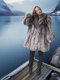 Person wearing a fur coat standing on a dock with a mountainous landscape in the background