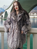 Woman wearing a fur coat standing by a waterfront with mountains in the background