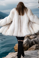 Person wearing a white fur coat standing on rocks by a body of water with mountains in the background.