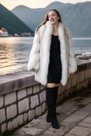 Woman in a white fur coat standing by a scenic lakeside with mountains in the background.