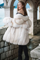 Woman wearing a white fur coat standing by a stone wall with a scenic background.