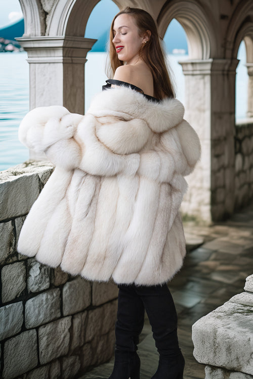 Woman wearing a white fur coat standing by a stone wall with a scenic background.