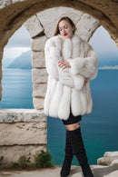 Woman in a white fur coat standing in front of a stone arch with a scenic background.