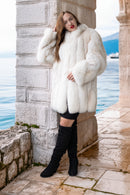 Woman wearing a white fur coat standing by a stone wall with water in the background