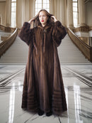 Woman in a long brown fur coat standing in an elegant interior setting.