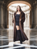 Woman in a long brown coat standing in an elegant hall with columns.