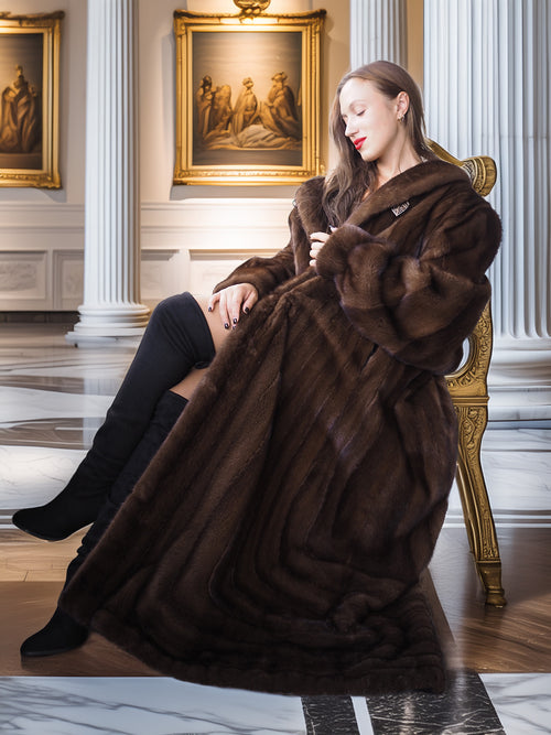 Woman in a brown fur coat sitting on a gold chair in an elegant interior setting.