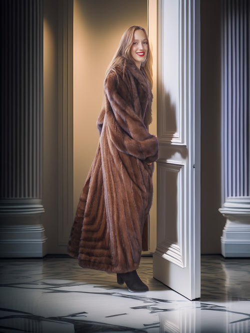 Woman in a long brown fur coat standing in a doorway with classical columns.