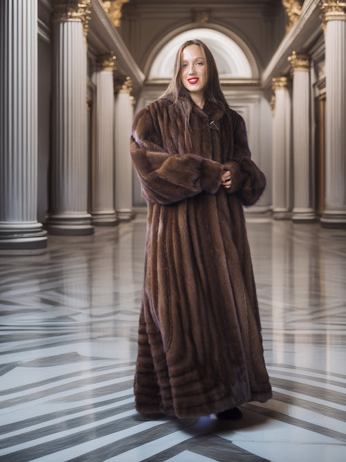 Woman in a long brown fur coat standing in an elegant hallway with columns.